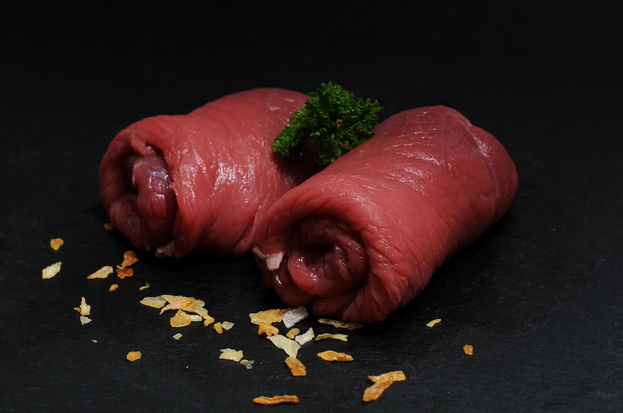 Involtini di carne dorati in teglia, ricoperti di salsa marrone, guarniti con prezzemolo fresco, disposti in file ordinata su fondo bianco