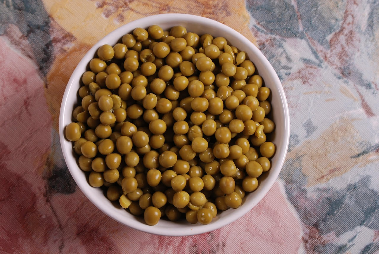 Un piatto di ceramica bianca contiene lagane larghe e ondulate di colore beige, condite con piselli verdi brillanti, una manciata di prezzemolo fresco tritato, e un filo di olio d'oliva. La pasta è cr