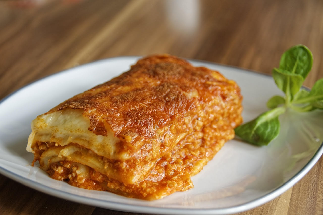Una porzione di lasagna bolognese tagliata mostra strati netti di pasta all'uovo gialla, ragù rosso-marrone e besciamella bianca cremosa, contorni leggermente dorati, piatto bianco