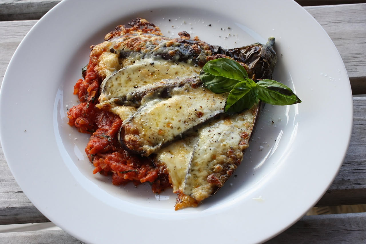 Melanzane alla cardinale in una pirofila di ceramica bianca, coperte di besciamella dorata e filamenti di formaggio gratinato al forno, vapore ancora visibile