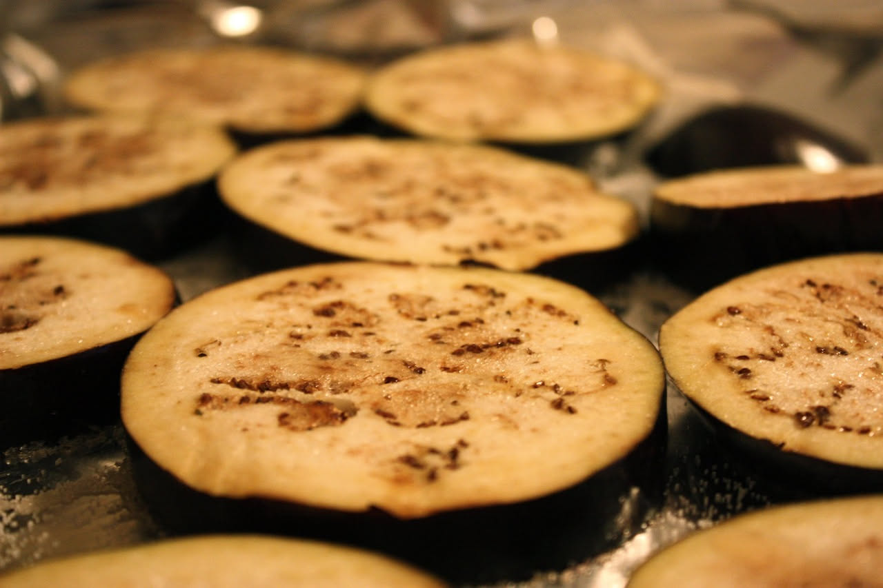 Fette di melanzana grigliata con segni del fuoco, disposte su un piatto bianco con fili di olio, aglio fresco e prezzemolo verde