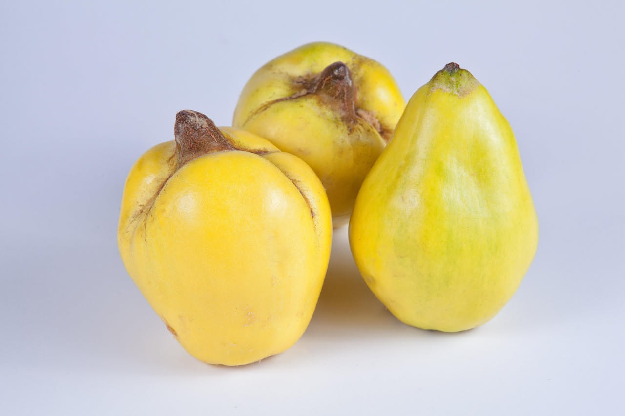 Vasetto di vetro trasparente con mele cotogne tagliate a dadini di colore giallo ambrato immersi in una salsa marrone scuro con semini di senape visibili sulla superficie, accanto a un piatto bianco c