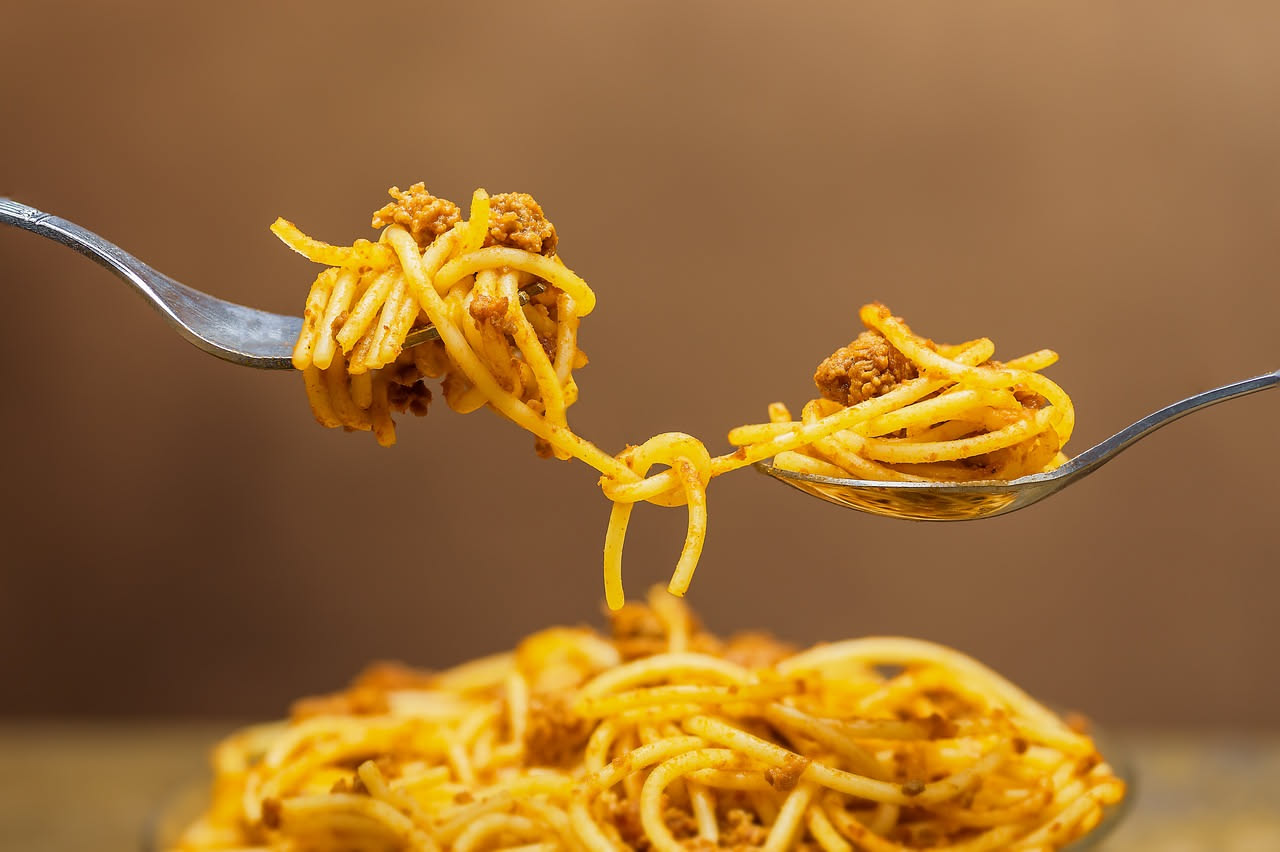 Piatto bianco di mezzani al ragù con sugo scuro e pezzetti di carne visibili, formaggio grattugiato sopra, forchetta d'argento di lato