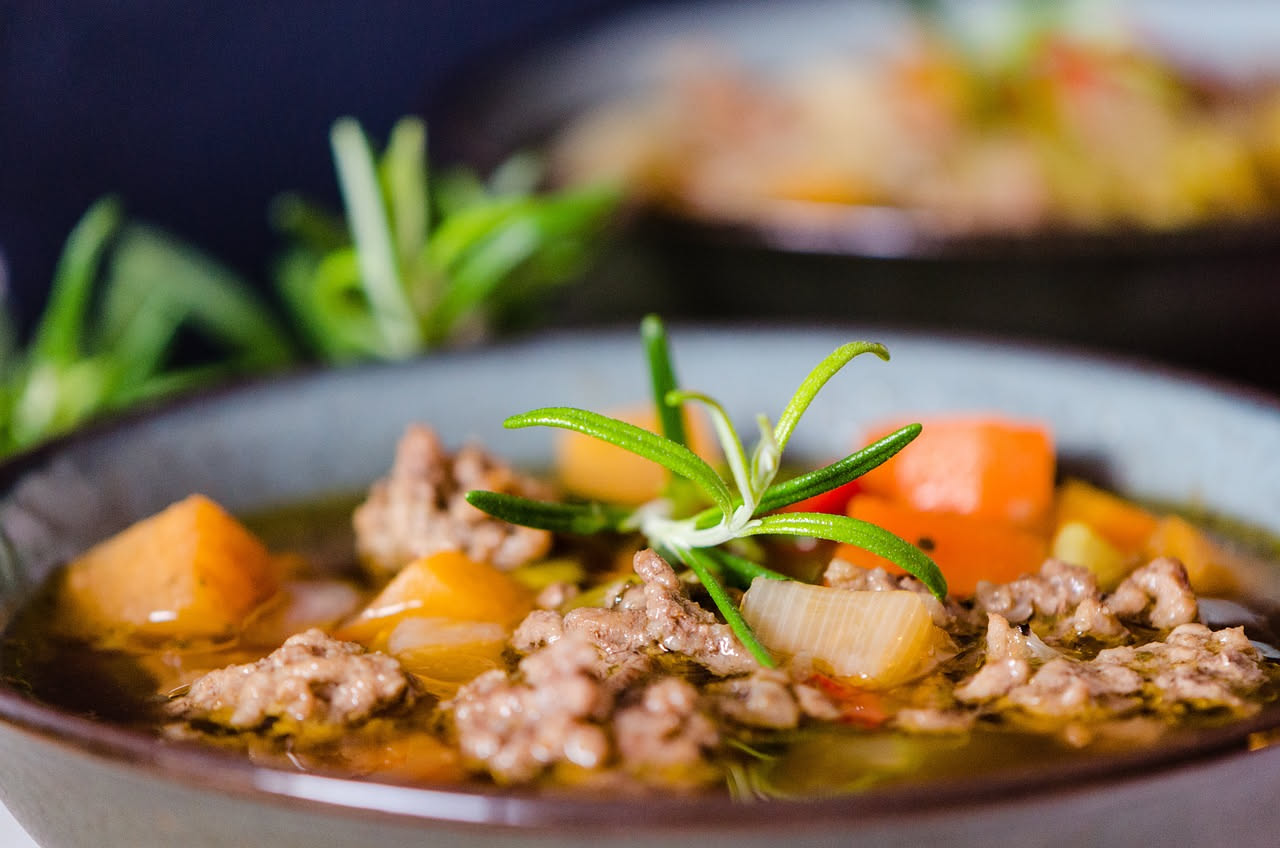 Una ciotola bianca con minestra d'orzata fumante: orzo gonfo e cremoso, verdure morbide, brodo trasparente dorato, guarnita con un filo di olio extravergine e prezzemolo fresco tritato