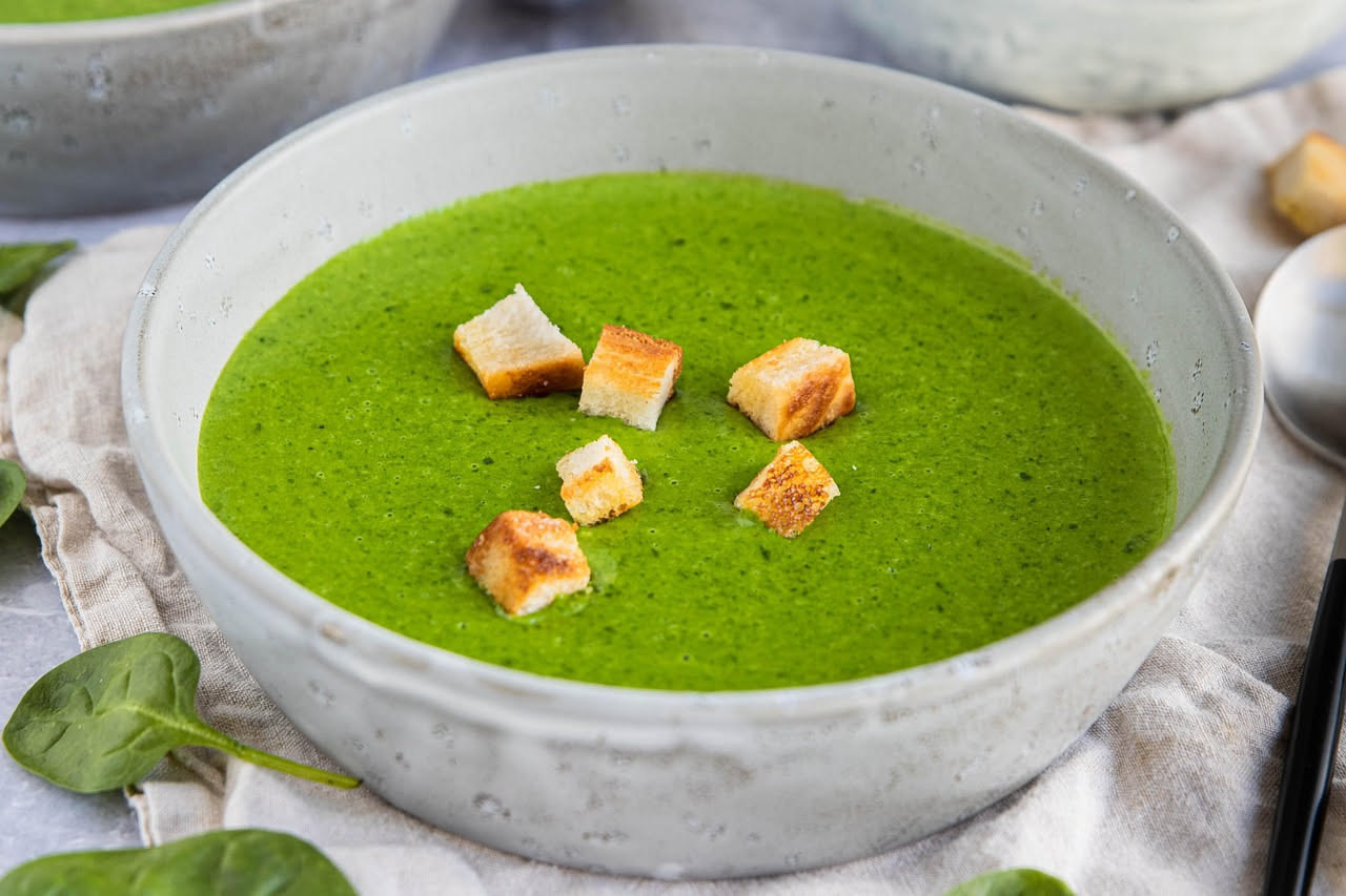 Una ciotola di ceramica bianca con minestra di cicoria: brodo trasparente con finiture dorate, cicoria verde scuro sopra, cubetti di pane tostato sparsi in superficie, cipolla morbida visibile, cucchi