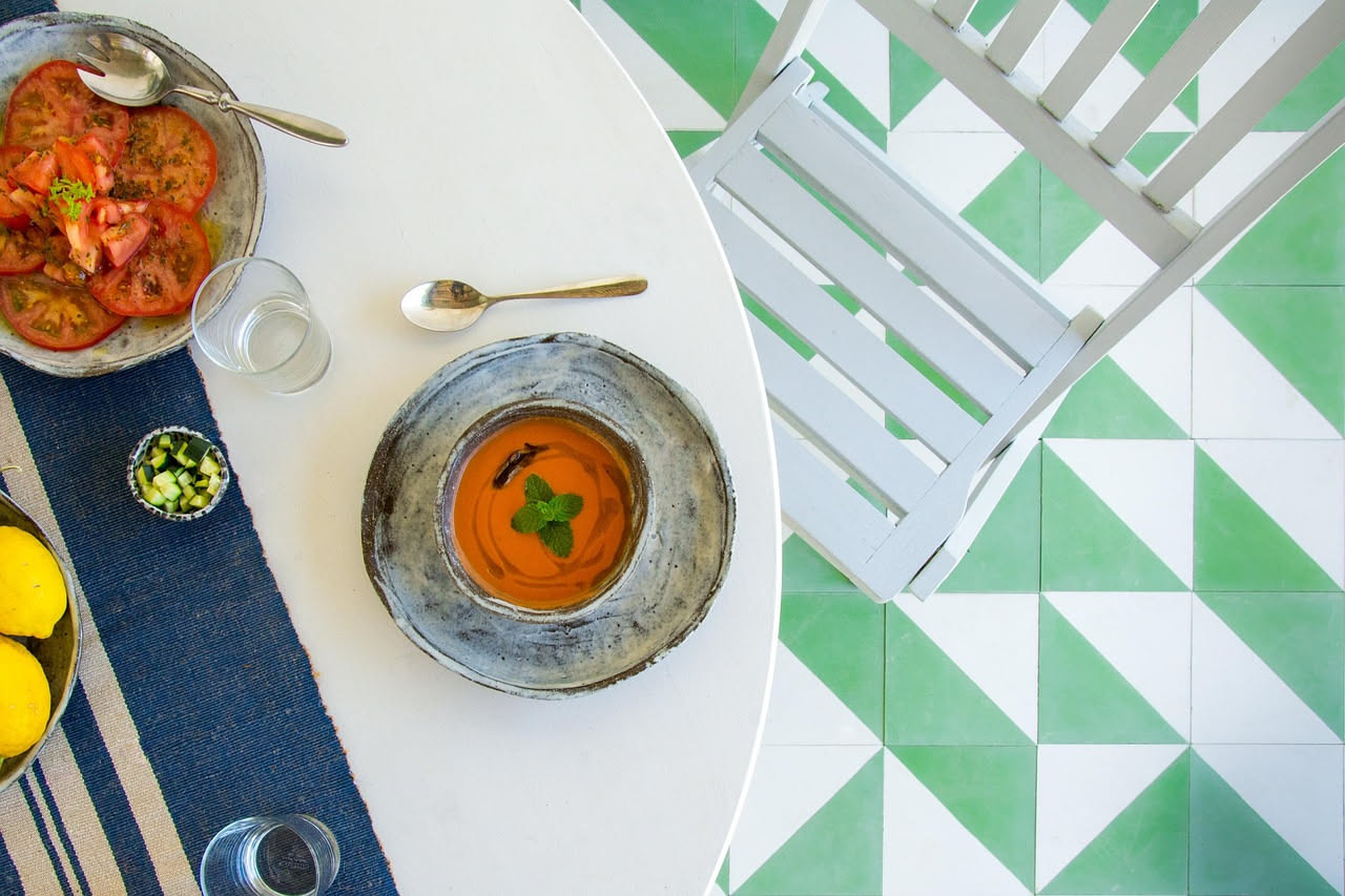 Una ciotola in ceramica bianca con minestra di fava beige-verde, cremosa, decorata con un filo di olio verde oliva e una foglia di menta fresca, su un tavolo di legno grigio con un cucchiaio ai lati.