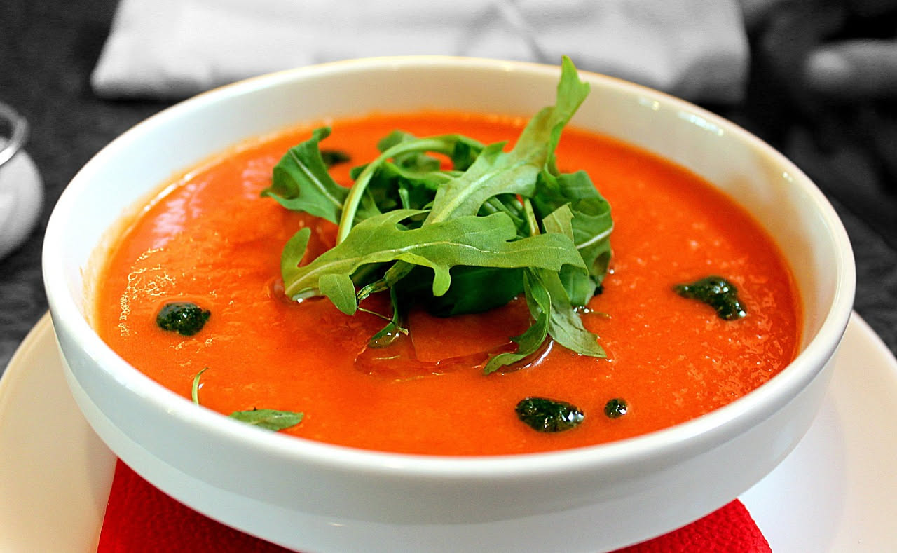 Ciotola di ceramica bianca con minestra di pappa al pomodoro di colore rosso-arancio, cremosa e densa, guarnita con foglie di basilico fresco e filo d'olio d'oliva verde, cucchiaio dentro il piatto, s