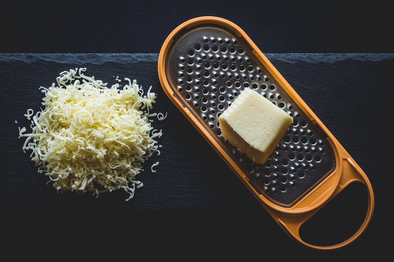Una forma di Montasio con crosta marrone dorato su tavolo di legno, accanto a un pezzo affettato che mostra la pasta giallo paglierino con occhiature, un coltello e una grattugia