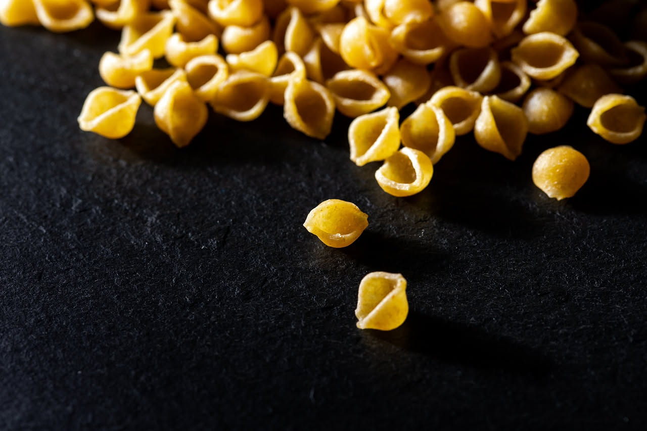 Mugnuli dorati e croccanti impilati su un piatto bianco, ricoperti di miele lucido, granella di pistacchio verde e mosto cotto scuro, con vapore leggero che sale