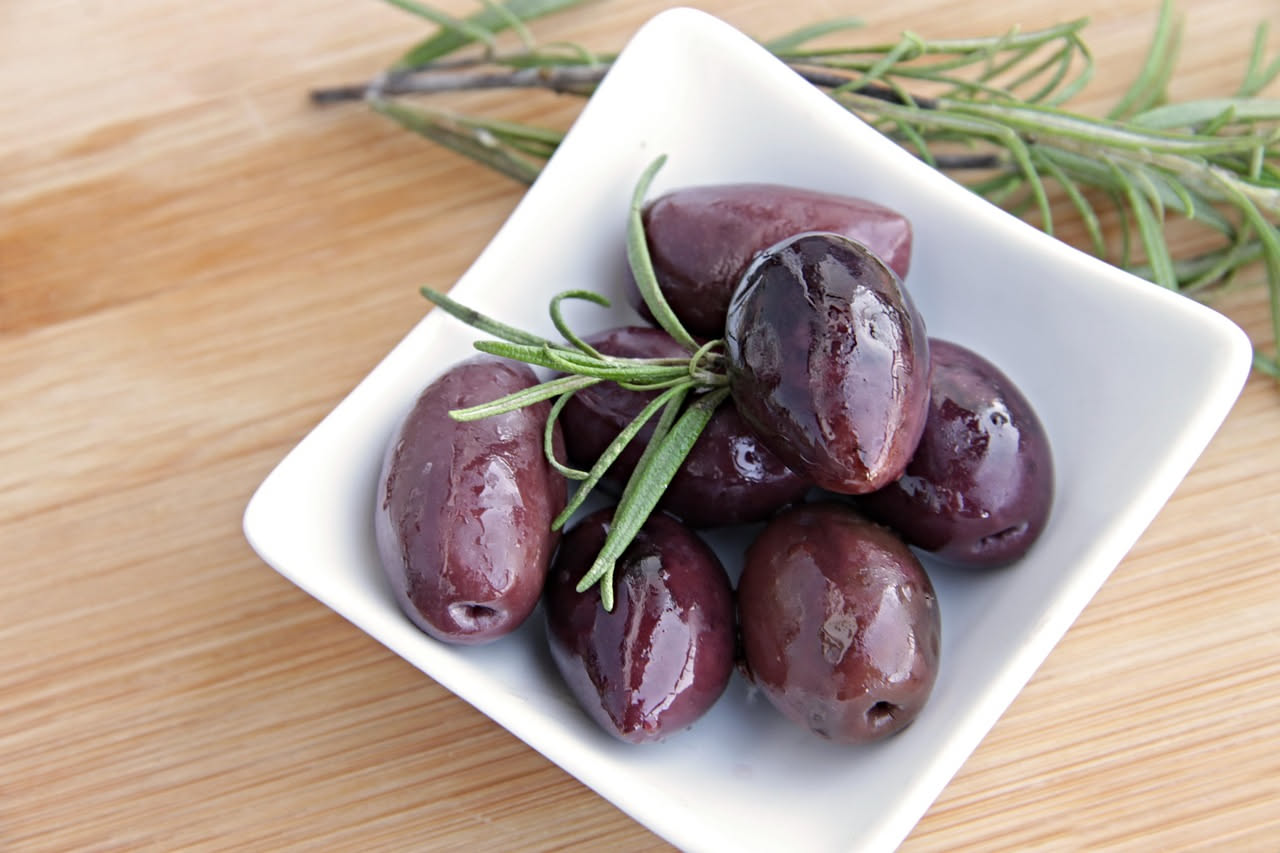 Barattolo di vetro trasparente con olive nere intere immerse in salamoia limpida, con rametti di rosmarino e spicchi d'aglio visibili nel liquido, fondo sfocato bianco