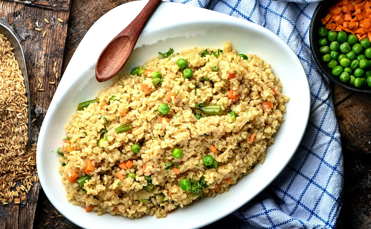 Piatto fondo bianco con orzotto cremoso color nocciola, zucchine verdi, carote arancioni e piselli sparsi in superficie, grattugia di parmigiano e basilico fresco
