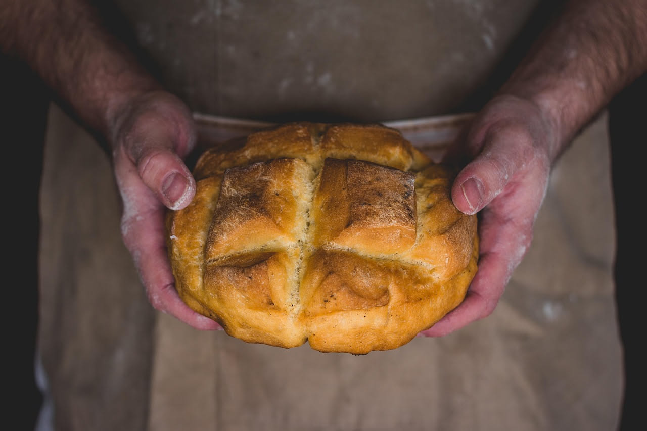 Pagnotta di pane ai ceci intera con crosta dorata e uniforme, tagliata a fette da cui si vede l'interno beige chiaro e soffice con piccoli buchi diffusi, posata su tavolo di legno con spighe di grano