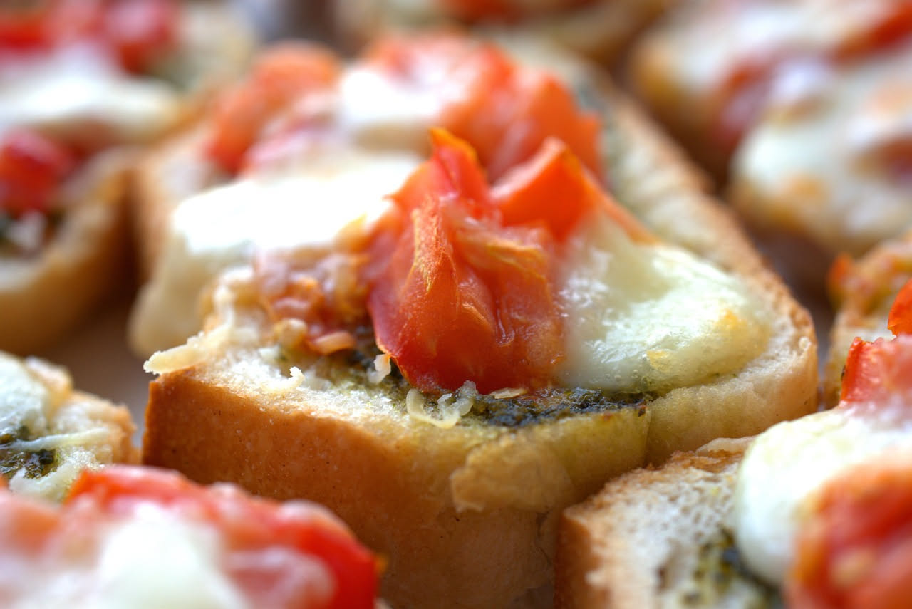 Tre fette di pane tostato su un piatto bianco, condite con pomodoro rosso tagliato, aglio affettato sottilmente, olio giallo-verde e basilico verde fresco