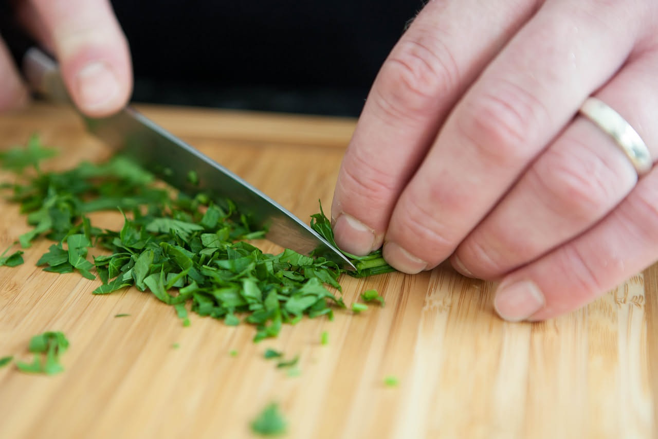Pappardelle larghe al prezzemolo in un piatto bianco fondo, condite con prezzemolo verde brillante tritato, visibili piccoli pezzi di aglio biondo, gocce di burro lucido, grattugia di parmigiano reggi