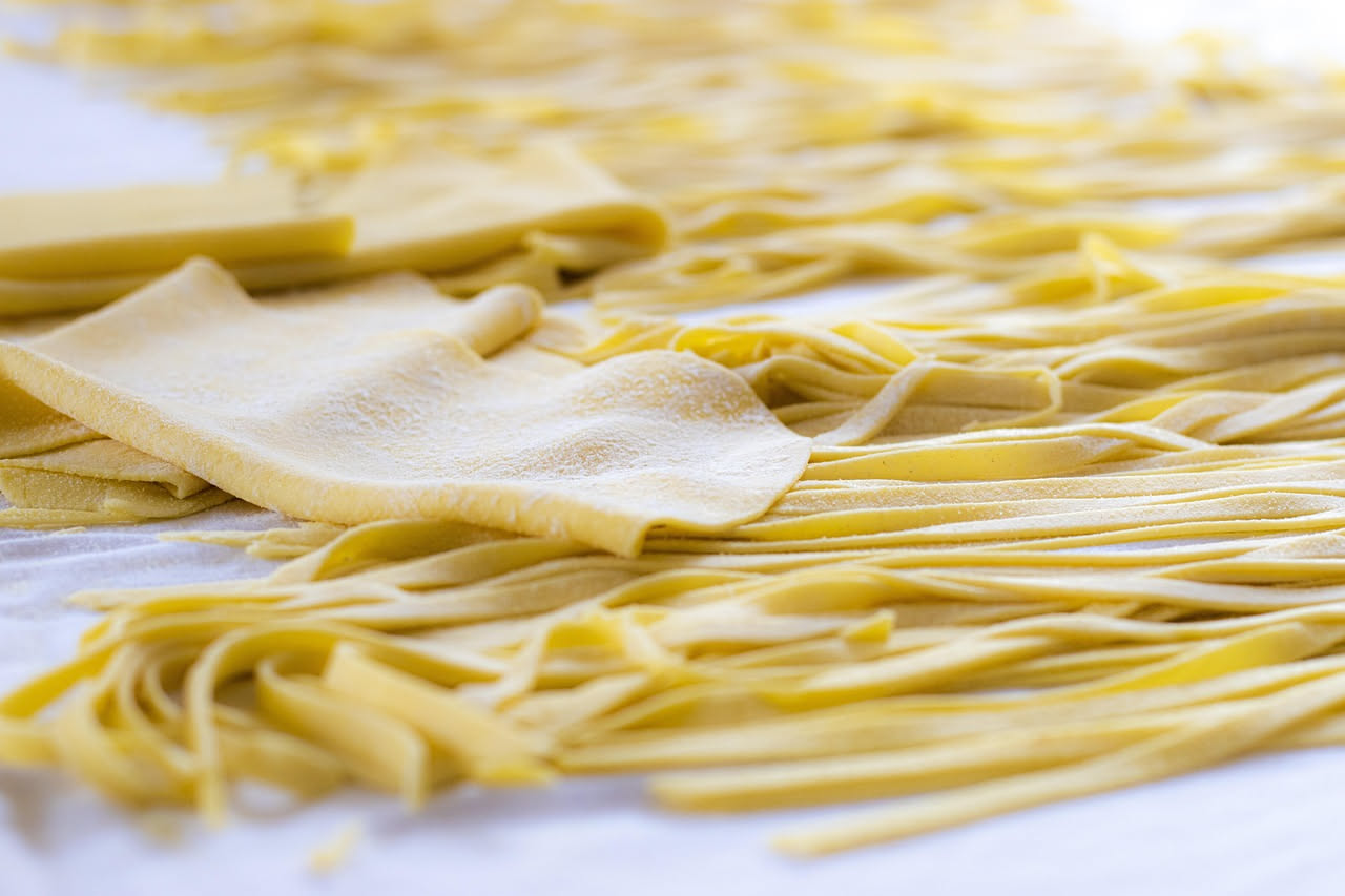 Pappardelle larghe gialle distese su piatto bianco coperte di ragù di lepre marrone scuro e denso, completate da grana padano grattugiato e prezzemolo verde spezzettato, impiattamento rustico