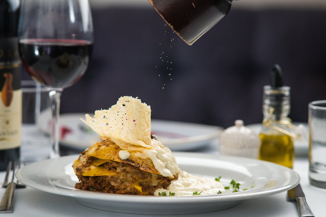 Scaglie sottili e ricurve di parmigiano reggiano bianco-crema posate su un piatto bianco, con riflessi lucidi, accanto a pepe nero grossolano e un coltello metallico.
