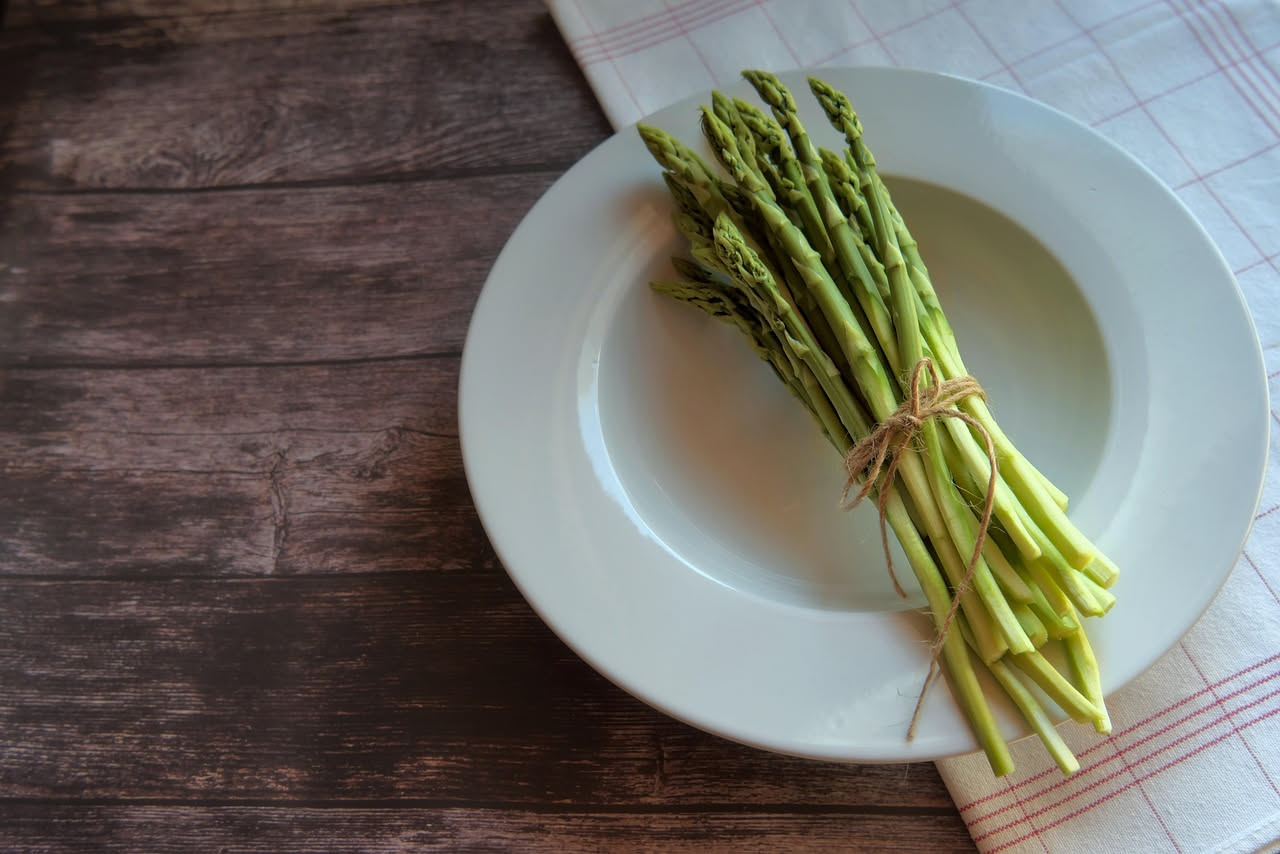 Piatto di pasta lunga con asparagi verdi teneri, condita con olio e grana, grani di pepe nero visibili, su fondo bianco