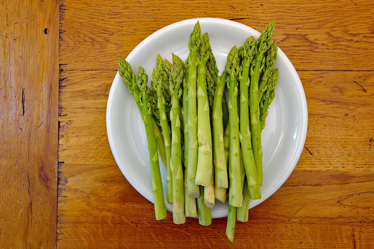 Un piatto di pasta bianca con asparagi verdi interi distribuiti sopra e intorno, granello di pepe nero visibile, olio che brilla sulla superficie, servito su piatto bianco con bordo sottile