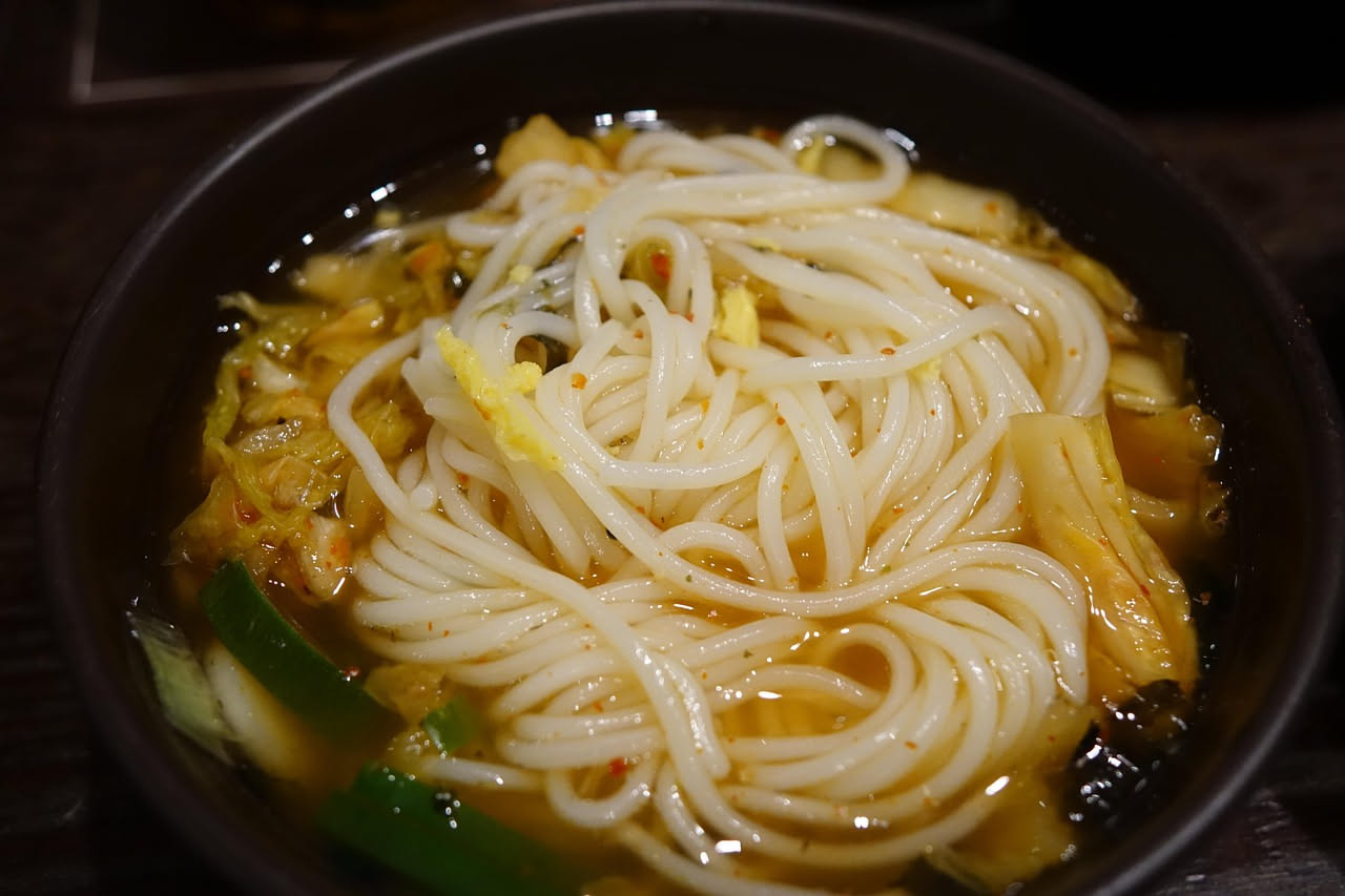 Ciotola di ceramica bianca con pasta e vermicelli immersi in brodo dorato trasparente, guarnita con una spruzzata di prezzemolo fresco, cucchiaio di legno appoggiato sul bordo