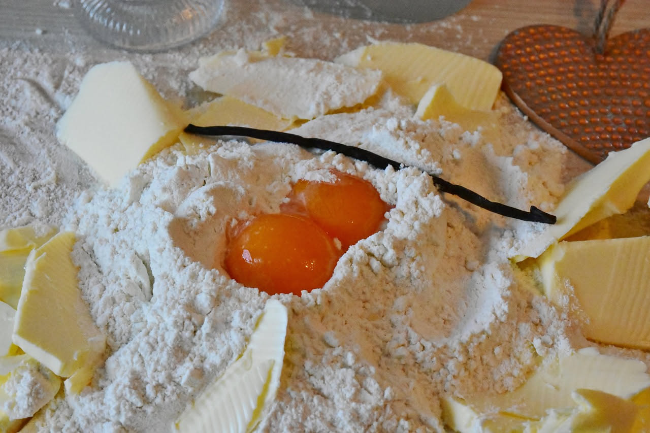 Sfoglia di pasta fresca giallo oro semitrasparente stesa su tavolo di legno bianco, con matterello affiancato e uova crude in ciotola di ceramica sullo sfondo