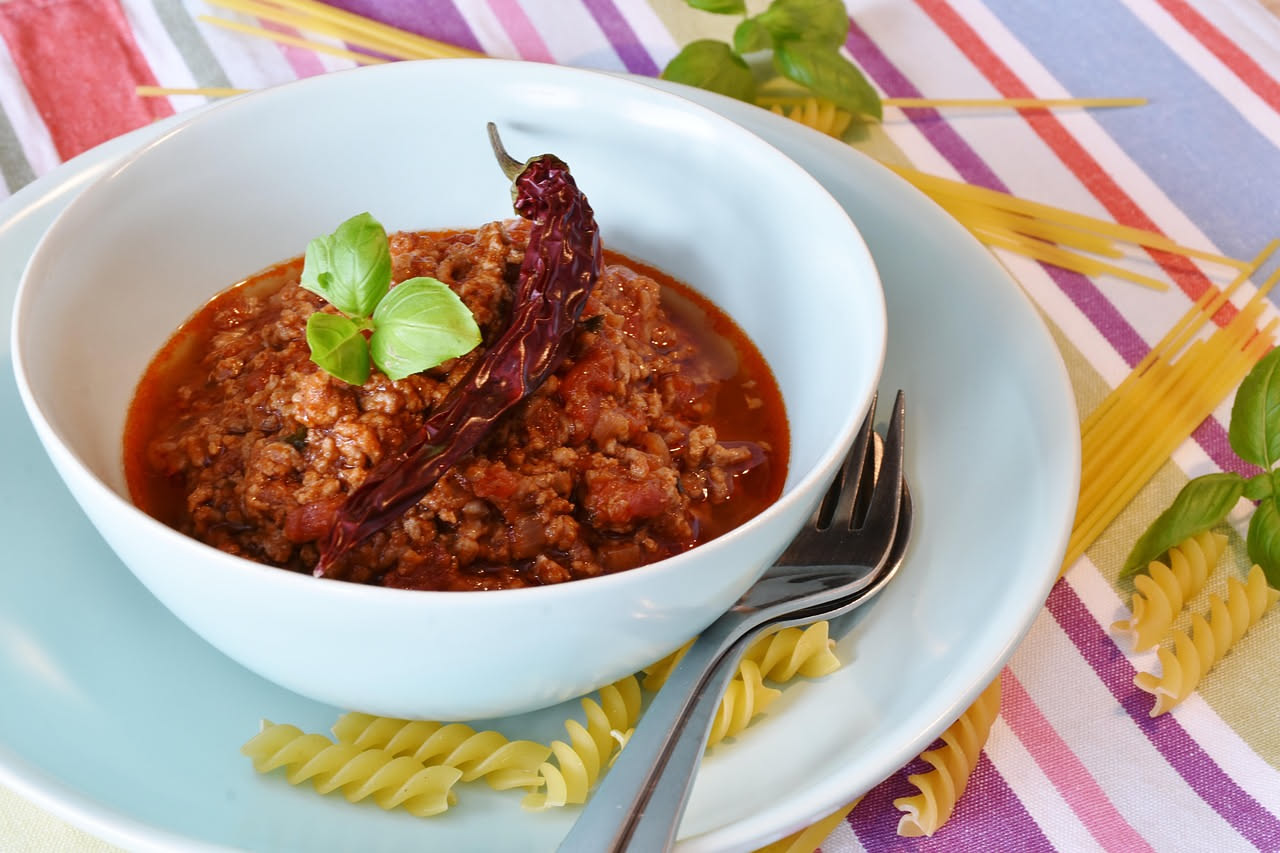 Un piatto fondo di ceramica bianca contiene brodo scuro rosso e un pezzo di carne marrone tenera circondata da cavoli e sedano; pasta fusilli al dente mista nel brodo; accanto un cucchiaio di legno e
