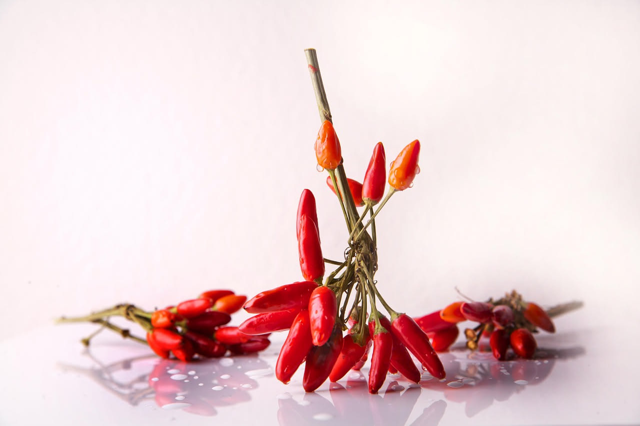 Vasetto di vetro trasparente con peperoncini rossi e verdi interi immersi in olio dorato, aglio sbucciato e foglie di prezzemolo fresco visibili tra i peperoncini