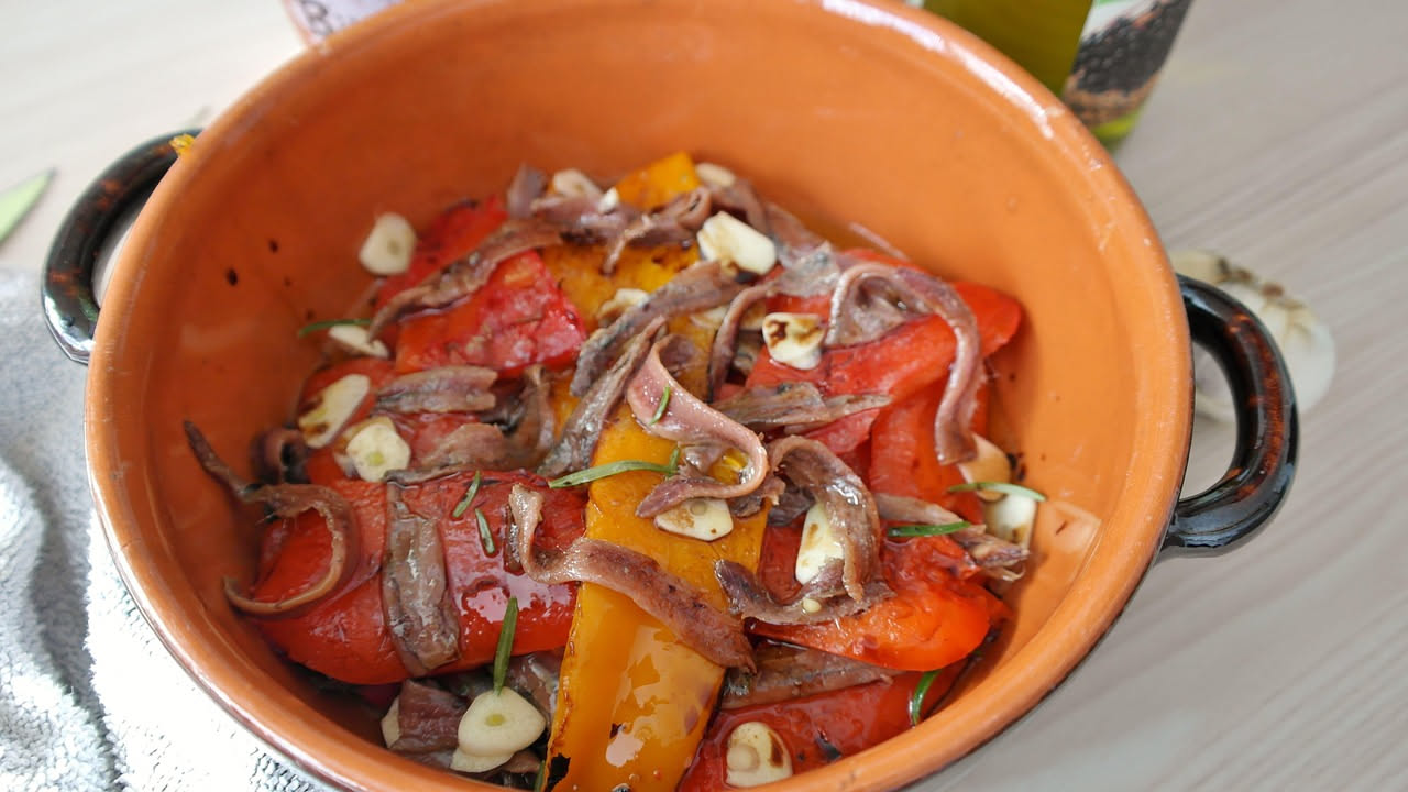 Peperoni rossi e gialli tagliati a strisce conditi con filetti di acciughe, olio d'oliva e aglio, in un piatto bianco