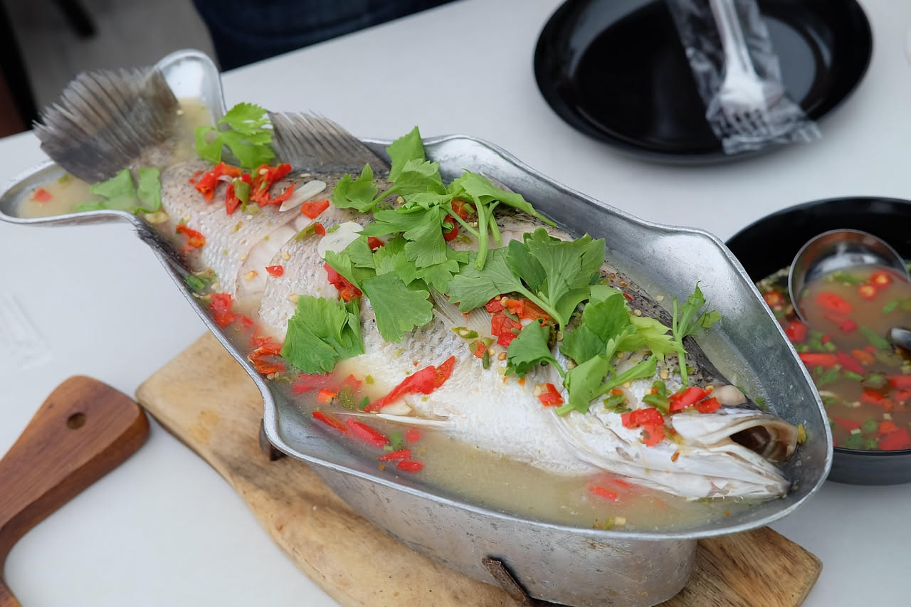 Un piatto fondo di ceramica bianca con persico intero cotto in brodo trasparente e fumante, guarnito con fettine di carota e sedano, prezzemolo fresco verde in superficie