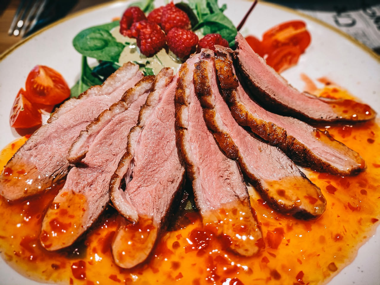 Petto d'anatra affettato nel piatto: fette di carne rosata circondate da pelle dorata e croccante, accompagnate da verdure scure di contorno e salsa marrone nel fondo del piatto bianco