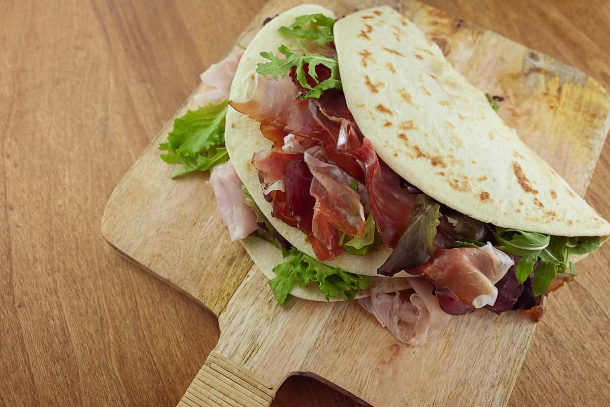 Piadina riminese appena cotta in padella, colore dorato e croccante, farcita con prosciutto crudo e squacquerone, su tavolo di legno