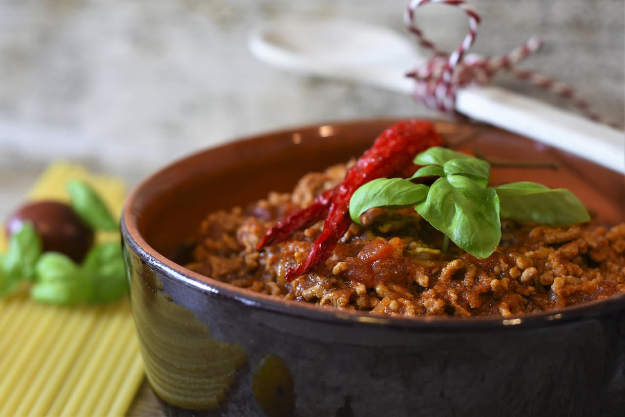 Piatto fondo con polenta giallo oro cremosa, coperta da ragù rosso scuro con pezzetti di carne, grattugia di formaggio bianco sulla superficie, cucchiaio di legno appoggiato al bordo
