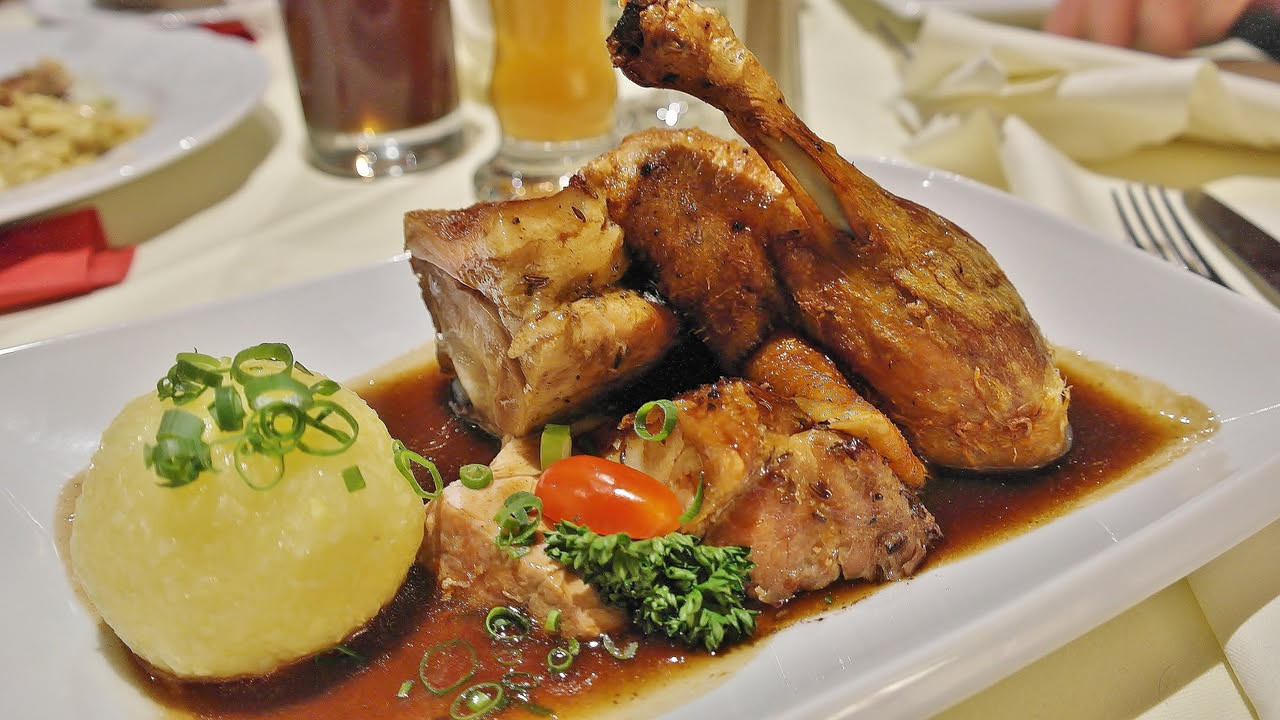 Pollo dorato con pelle rosso-marrone brillante in una cocotta di terracotta con salsa scura e cipolla caramellata, vapore appena accennato, contorno di patate sul piatto bianco