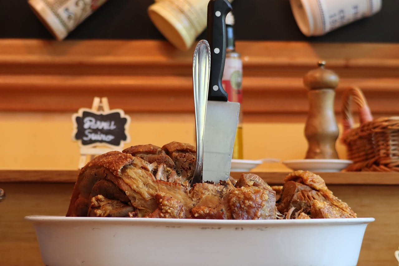 Porchetta intera cotta al forno dorata e croccante, affettata su un vassoio di legno, circondata da rosmarino e alloro, con la pelle lucida e caramellata