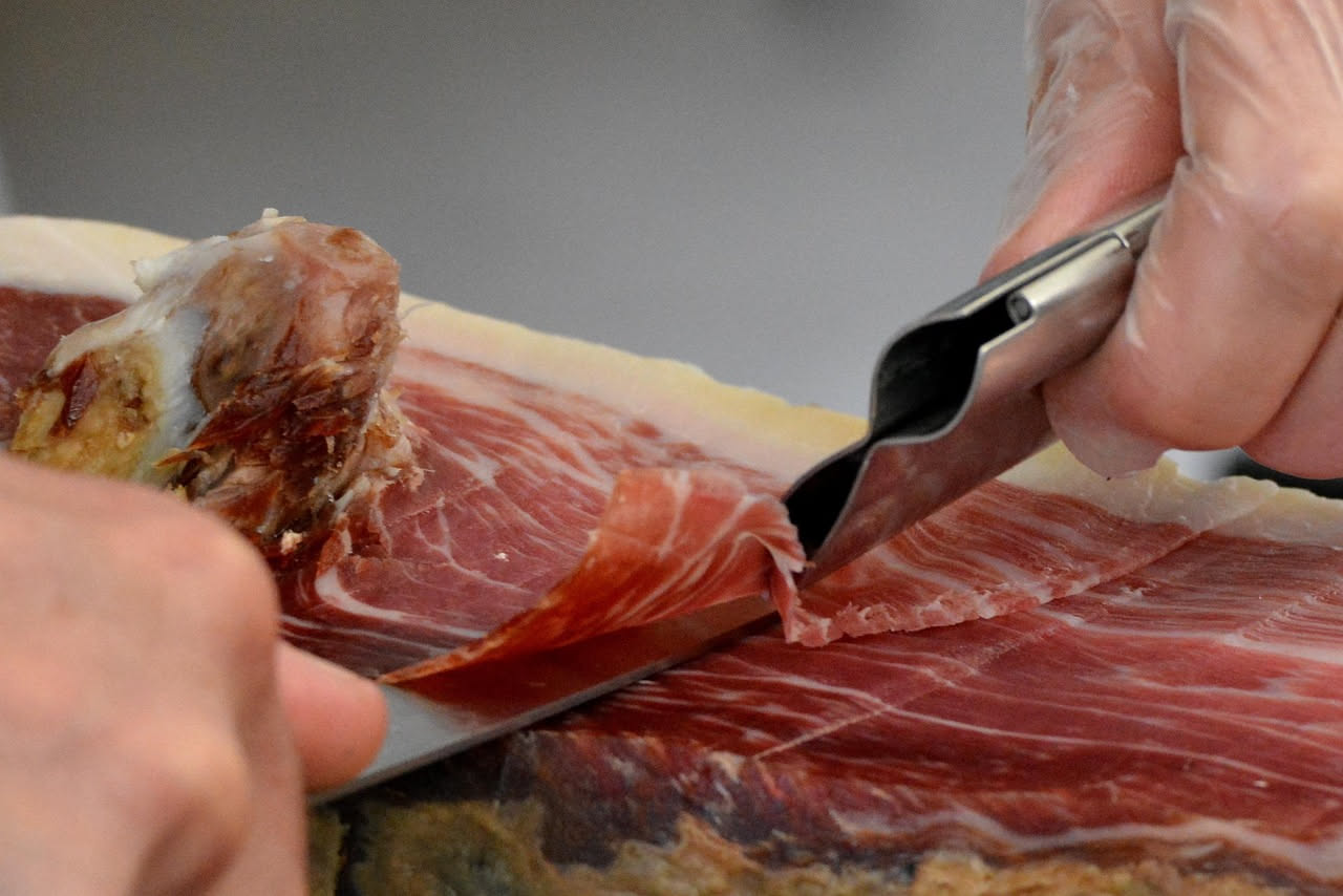 Fette trasparenti di prosciutto di Parma rosa pallido disposte a ventaglio su un piatto bianco, con un coltello da affettatura accanto