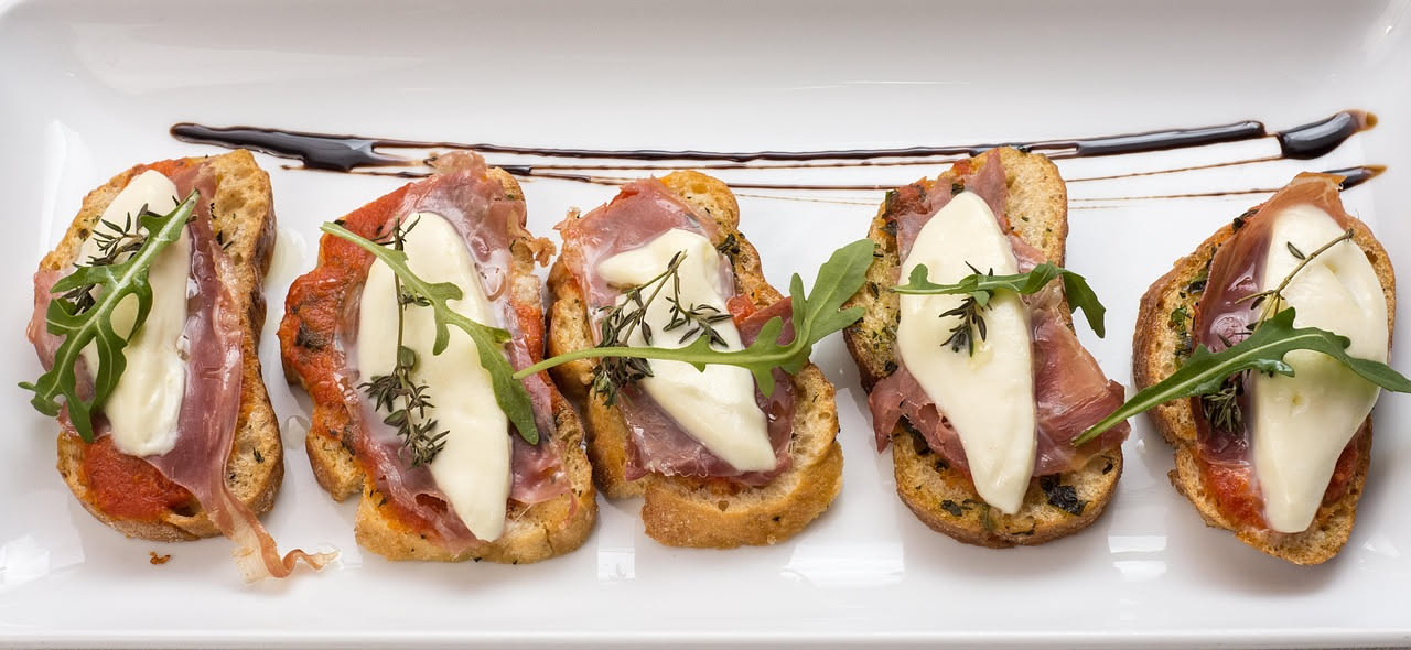 Crostini di pane toscano tostato con fette sottili di prosciutto toscano rosso scuro, un filo d'olio d'oliva in superficie, su un piatto bianco