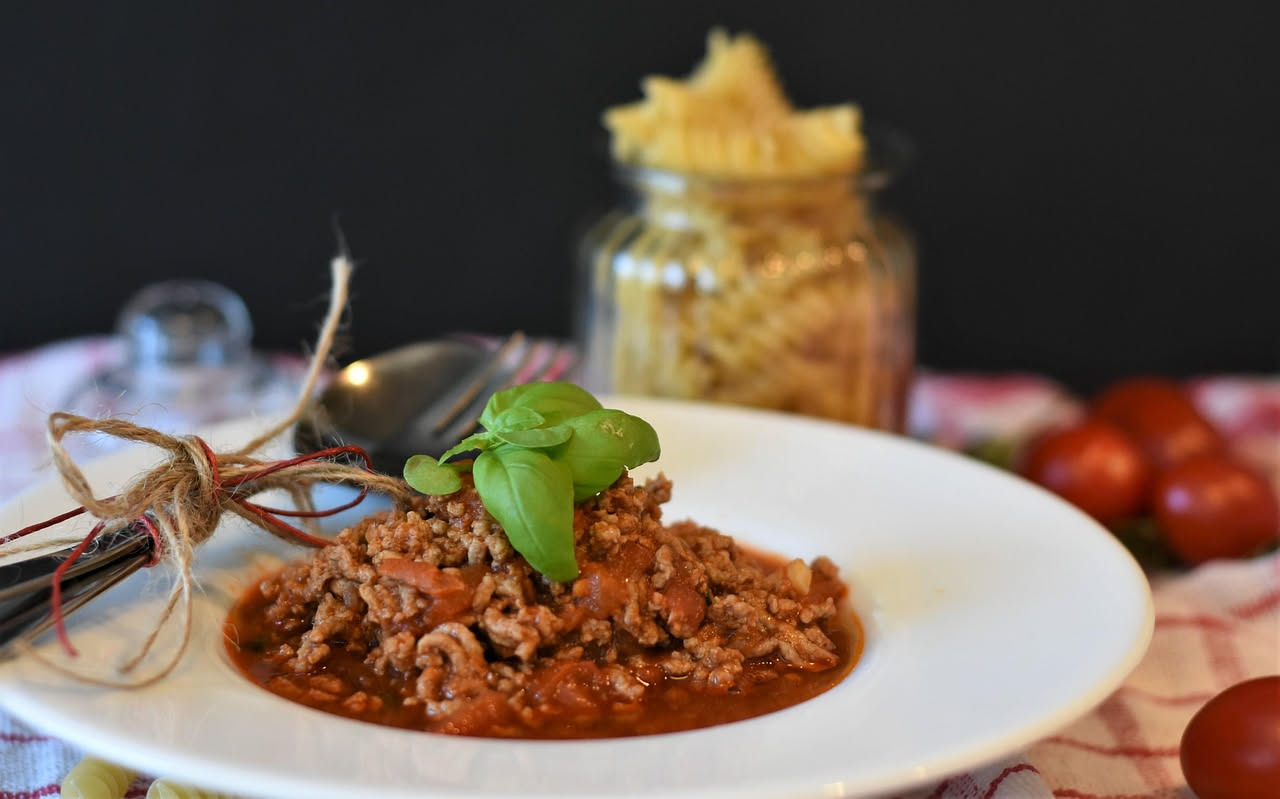 Piatto bianco con pasta fredda condita con ragù freddo rosso scuro, accanto un bicchiere di acqua fresca e foglie di basilico fresco verde, tavolo di legno naturale