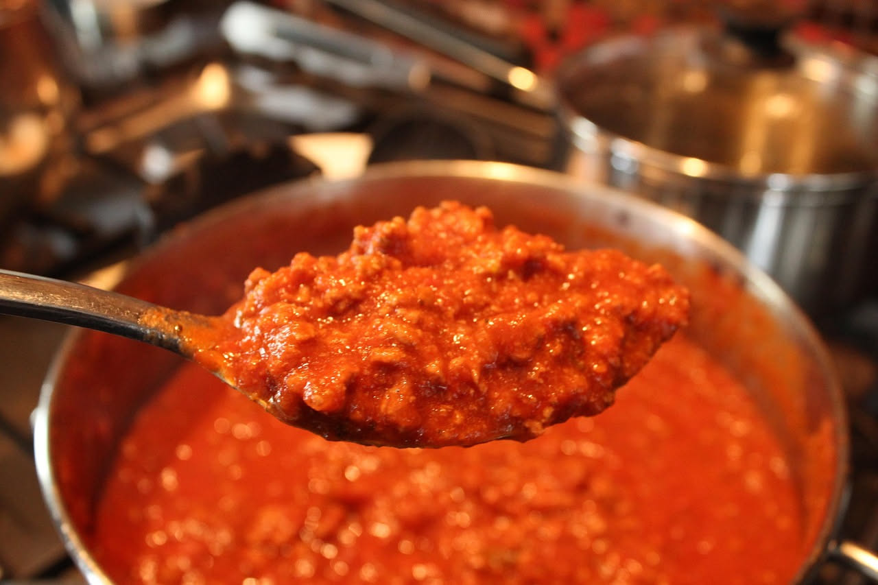 Una ciotola di ragù di carne mista color marrone scuro, denso e cremoso, servito in piatto bianco con sfumature di rosso dai pomodori, con spaghetti arrotolati in primo piano