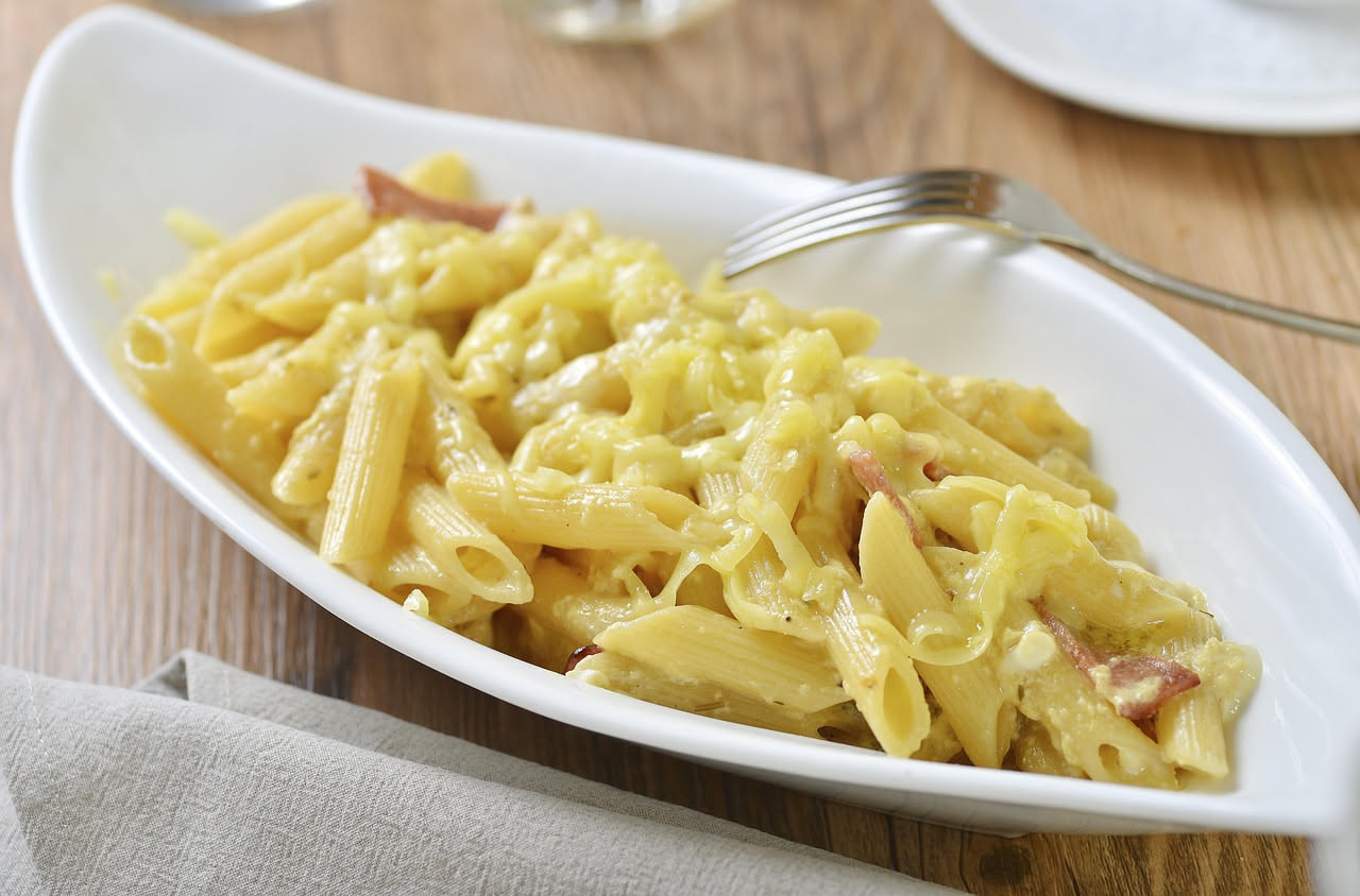 Piatto di rigatoni bianchi con condimento di guanciale croccante, formaggio pecorino giallognolo e pepe nero visibile, in ciotola di ceramica bianca, con grana pecorino e pepe accanto
