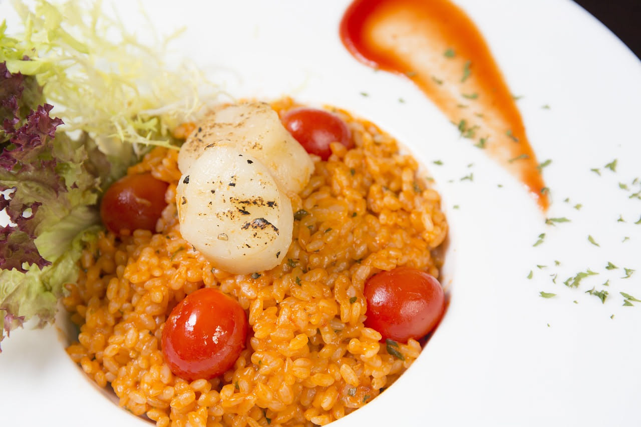 Piatto bianco con risotto cremoso di colore bianco avorio, guarnito con uova di riccio di mare di colore arancione intenso, filo di olio extravergine lucido in superficie, sfondo neutro