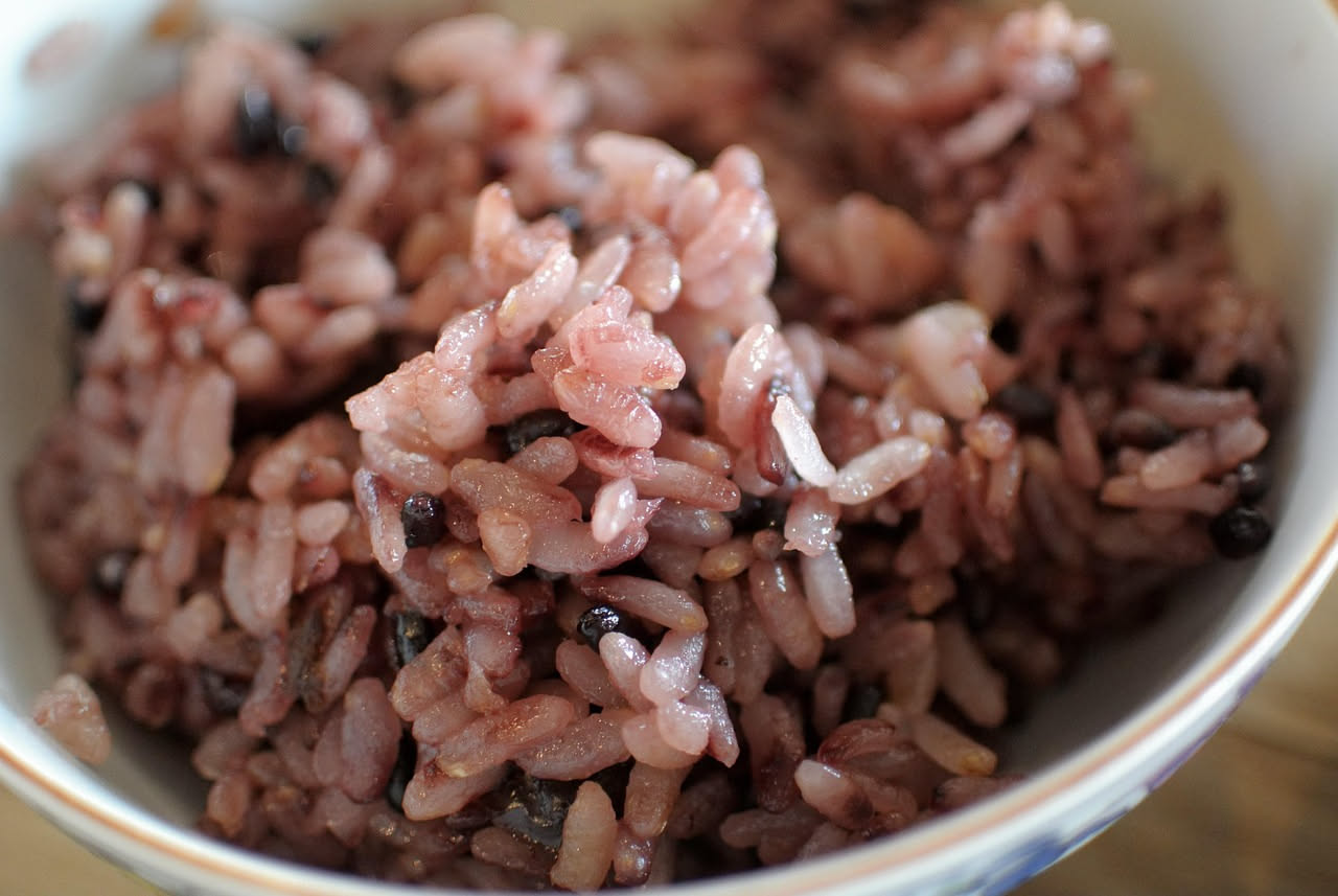 Piatto di risotto al Lambrusco servito in ciotola bianca, colore giallo cremoso con riflessi rosa delicati dal vino, mantecatura visibile, burro e formaggio grattugiato in superficie, decoro di prezze