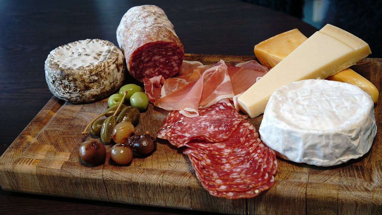Salame d'oca affettato su un tagliere di legno, con fette rotonde color rosso-marrone, tra acini d'uva nera e pane tostato. Sfondo neutro con luce naturale.