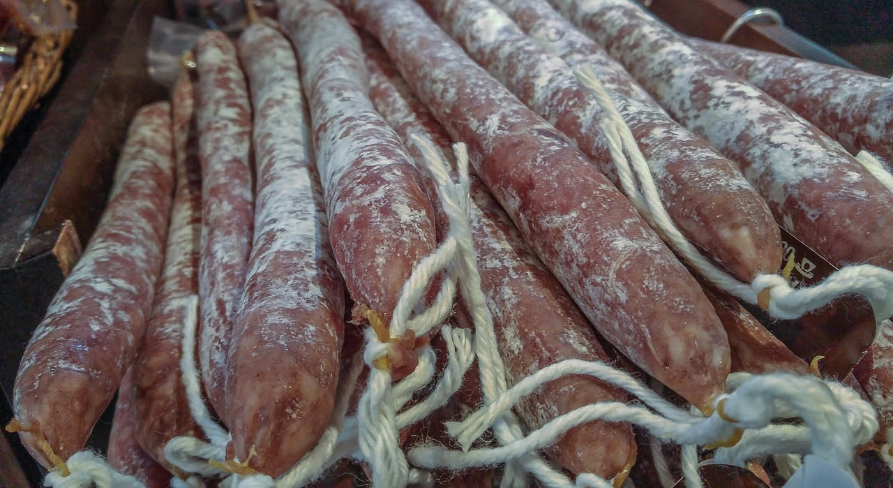 Salame della duja affettato su un piatto bianco, con fette trasparenti di grasso dorato intorno, coltello da affettare e una ciotola di senape in sottofondo