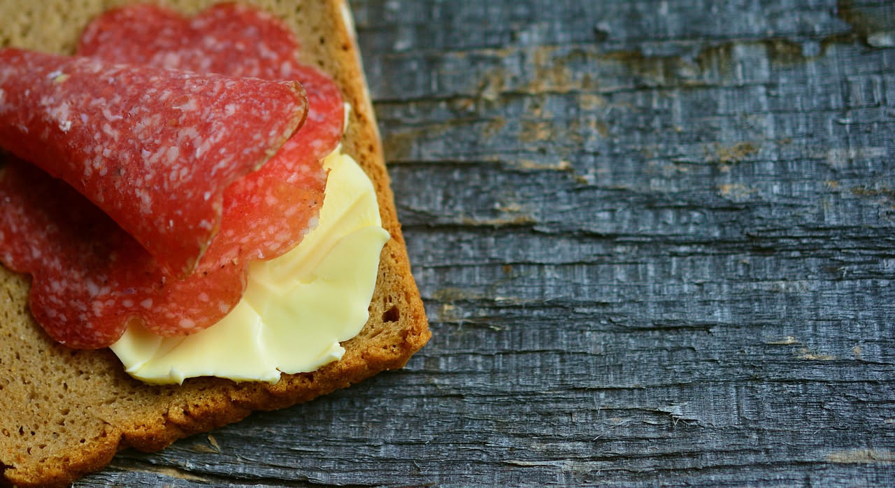 Salame gentile affettato su un tagliere di legno naturale, con una trama omogenea rosa pallido e spezie visibili, accanto a pane tostato e un bicchiere di vino bianco
