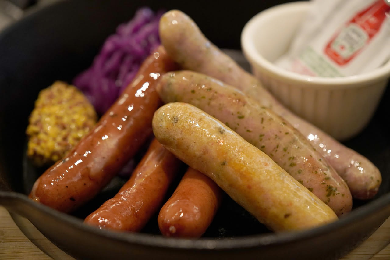 Tre salsicce cotte dorate e succose in una padella di ghisa, circondate da peperoni gialli e rossi a striscie, contorno di erbe aromatiche fresche appena colto