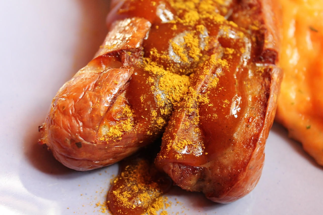 Salsiccia di Bassano cotta in padella, di colore marrone dorato con la superficie caramellata, accompagnata da fettine di cipolla e un rametto di rosmarino fresco