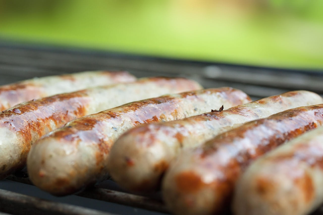 Tre salsicce fresche di montagna gonfie e dorate, crude su un tagliere di legno con aglio, rosmarino e pepe nero sparsi intorno