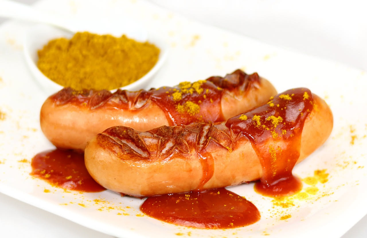 Salsicce marroni e lucide in un piatto fondo bianco, immerse in sugo rosso con pomodori, contornate da cipolla dorata, con prezzemolo fresco sparso sopra