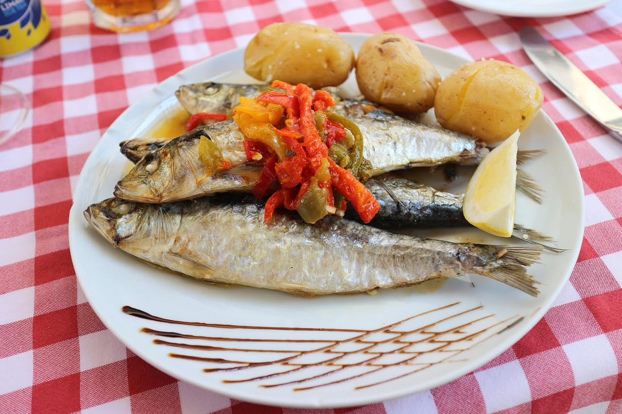 Sardine dorate fritte disposte in un piatto bianco coperte di salsa agrodolce con cipolla bianca affettata e pinoli, con aceto visibile sulla superficie, contorno di verdure grigie sfuocate