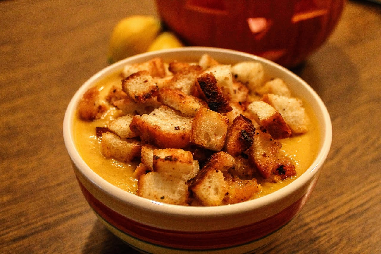 Ciotola bianca con sopa di pane fumante: pane biancastro in brodo denso, con verdure verdi e arancioni visibili, orlo d'olio dorato in superficie, seduto su tavolo di legno chiaro