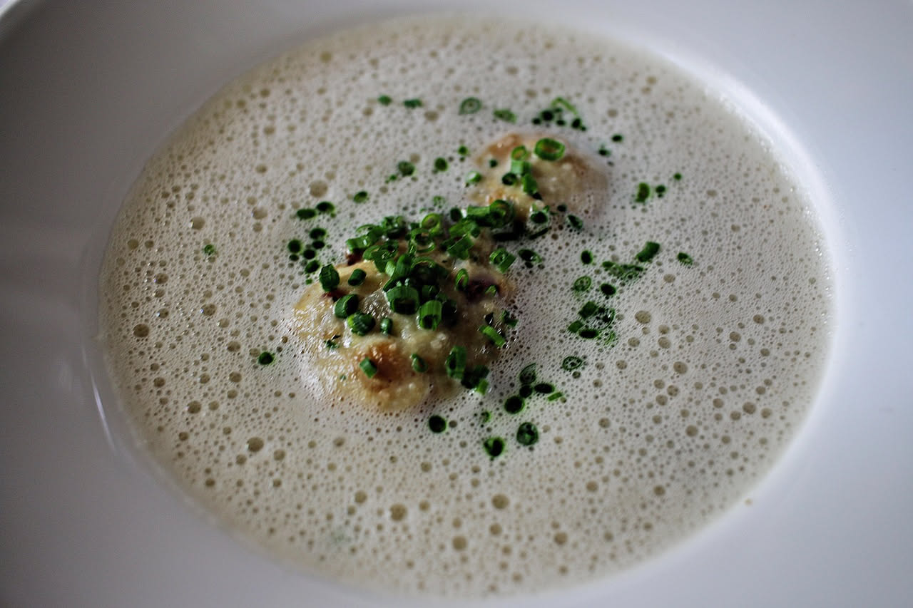Ciotola di ceramica beige con zuppa di cipolle color ambra trasparente, pane tostato galleggiante, formaggio fuso dorato in superficie, vapore visibile, cucchiaio di metallo appoggiato al bordo